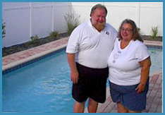 A man and woman standing together on the poolside patio of an in-ground swimming pool, smiling at the camera. Both are casually dressed in white tops and shorts. The pool area is surrounded by a white fence and some landscaping.