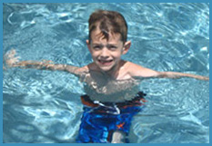 A young boy with short brown hair and blue swim trunks smiles while standing in a swimming pool with water up to his chest. His arms are outstretched, and he looks happy and playful in the clear blue water.