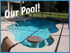 A backyard swimming pool with clear blue water, a curved design, and a submerged round table. "Our Pool!" is written in white text above the pool. In the background, there is a grassy yard with trees.