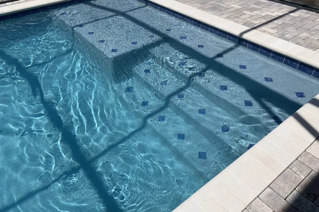 Close-up of pool steps with a glass tile accent band and a white paver deck